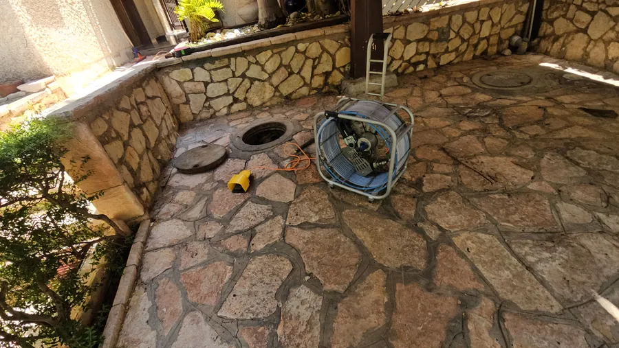 זירת עבודה לפתיחת סתימה בחצר מרוצפת עם שוחת ביוב פתוחה ומכונת שטיפה בלחץ