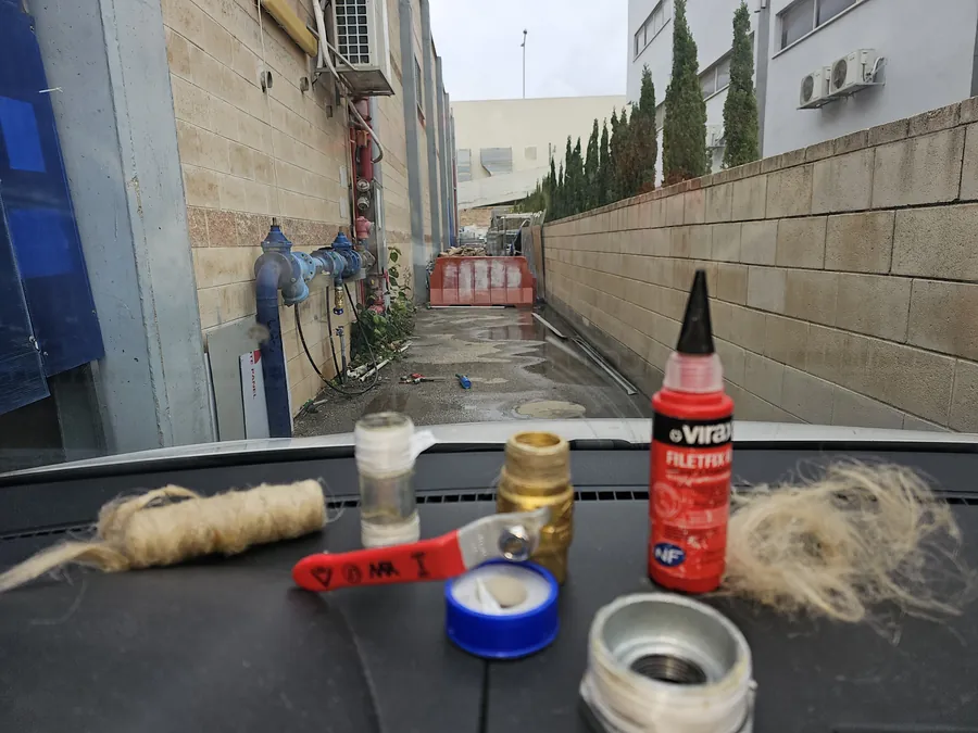 חומרי איטום לתיקון צנרת על לוח הרכב: פשתן, סרט טפלון, ברז כדורי ומשחת איטום, ברקע קו מים ראשי של בניין