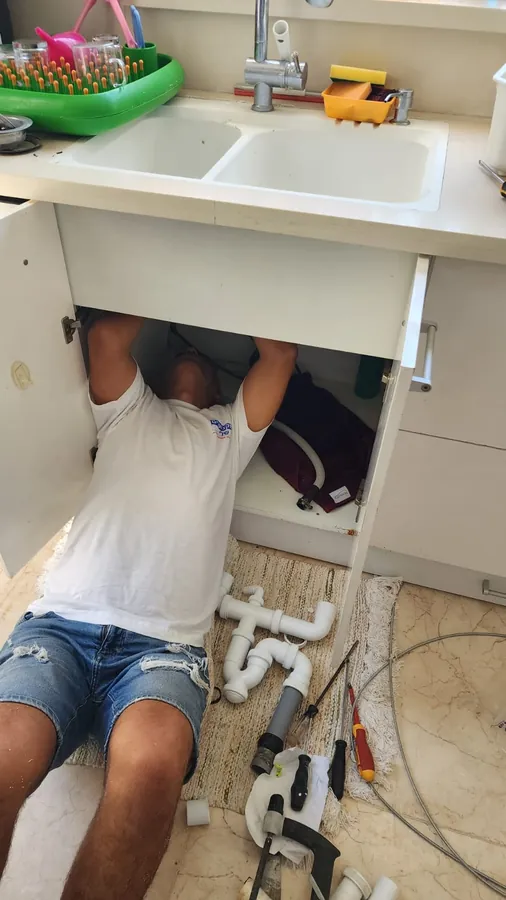 החלפת סיפון מתחת לכיור מטבח: האינסטלטור שוכב מתחת לארון והחלקים החדשים של הסיפון מונחים על הרצפה
