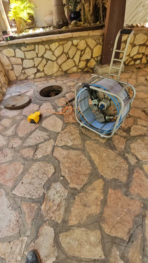 שוחת ביוב פתוחה בחצר מרוצפת לצד מכונת שטיפה בלחץ על גלגלים